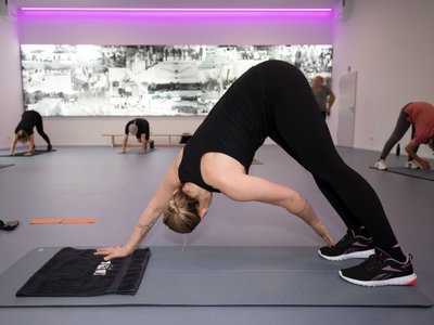 Bllick in einen Übungsraum. Eine Dozentin macht eine Gymnastikübung vor. Im Hintergrund sind Teilnehmende zu erkennenbung