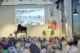 Das Bild blickt von hinten auf die Bühne zum Redner Thomas Beese. Seine Präsentation zeigt ein historisches Bild eines alten Bahnwaggons, aus den Fenstern winken Menschen.